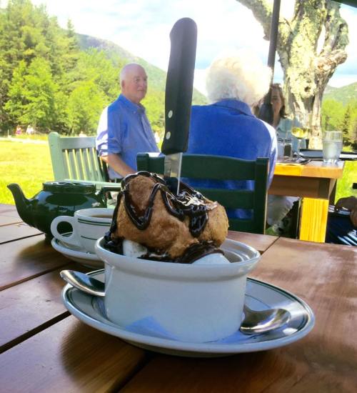 A dessert with ice cream and chocolate sauce sits on a table, with people and a scenic background in soft focus.