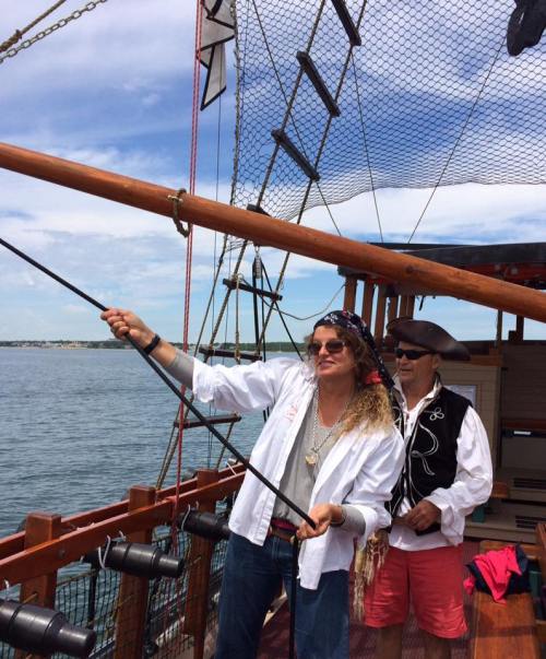 Two people on a boat: one in a pirate costume holding a fishing rod, the other in casual attire, enjoying the view.