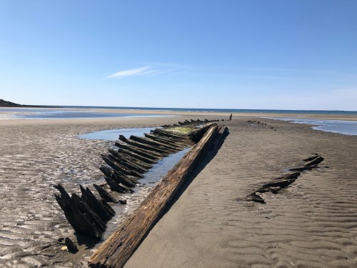 A weathered shipwreck partially buried in sandy beach, with calm water and clear blue sky in the background.