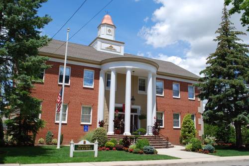 A brick building with a white columned entrance, surrounded by greenery and a blue sky with clouds.