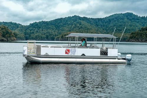 A pontoon boat with a canopy glides across a calm lake, surrounded by green hills under a cloudy sky.