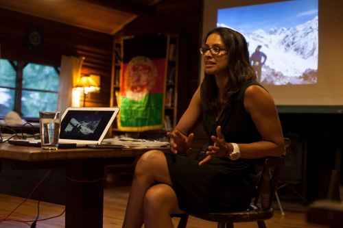 A woman in a black dress speaks passionately at a table, with a laptop and a mountain image projected behind her.