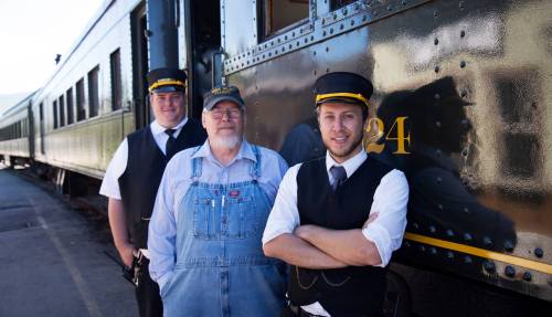 Three train crew members pose in front of a vintage train car, wearing uniforms and smiling at the camera.