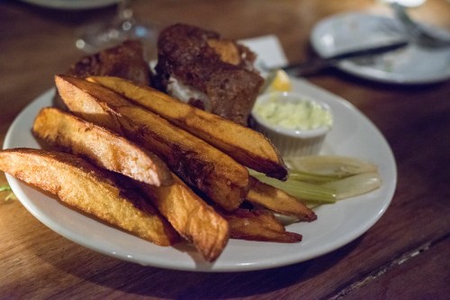 A plate of golden-brown fries with a side of dipping sauce and a piece of fried food, set on a wooden table.
