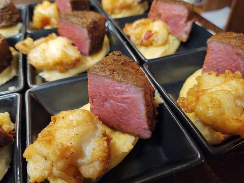 Plates of tender steak and shrimp served on creamy mashed potatoes, arranged in black square dishes.