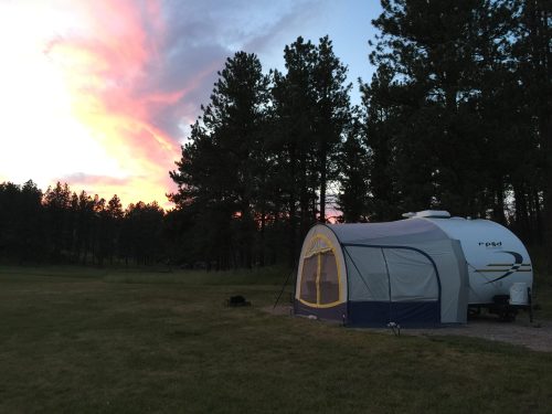 A camping scene featuring a trailer and a tent, surrounded by tall trees under a colorful sunset sky.