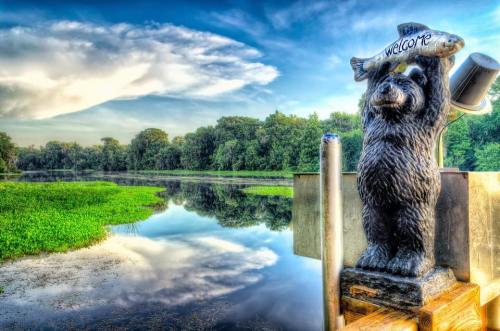 A bear statue holding a fish welcomes visitors by a serene lake surrounded by lush greenery and a blue sky.