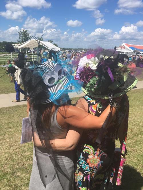Two women embrace, wearing colorful, elaborate hats adorned with flowers and decorations, against a sunny outdoor backdrop.