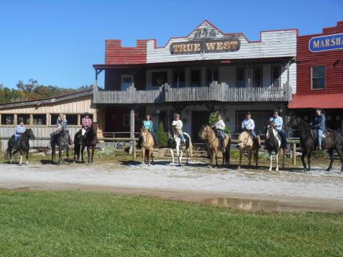 A group of people on horseback in front of a western-style building, with blue skies and green grass in the background.