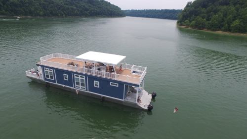 A blue houseboat floats on calm water, surrounded by green hills, with people enjoying the deck and swimming nearby.
