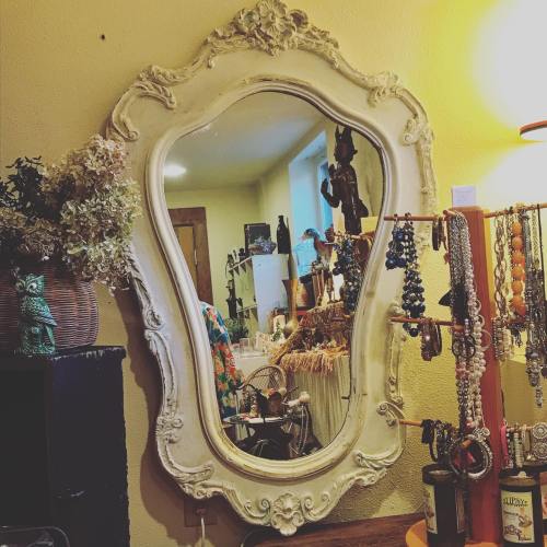 An ornate vintage mirror reflects a cozy shop filled with decorative items and jewelry.