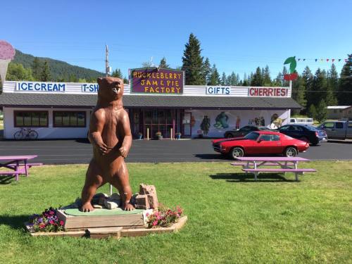 A wooden bear statue stands in front of the Huckleberry Jam Pie Factory, with colorful signs and picnic tables nearby.