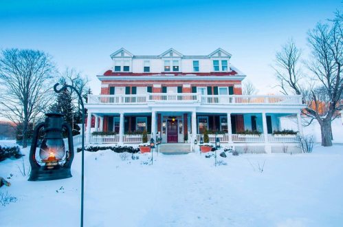 A large, historic house with a red roof, surrounded by snow, and a lantern in the foreground. Clear blue sky above.