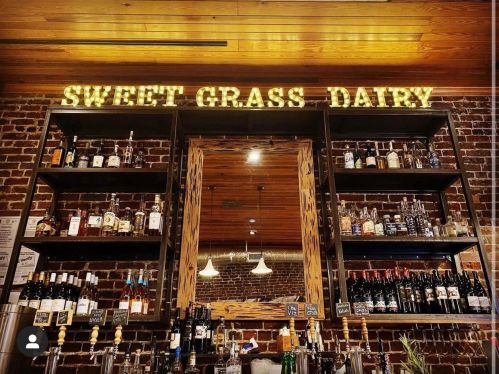 A rustic bar with shelves of various bottles and a sign reading "SWEET GRASS DAIRY" illuminated above.