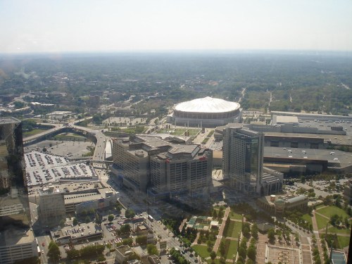 Aerial view of a cityscape featuring a stadium, parking lots, and surrounding buildings under a clear sky.