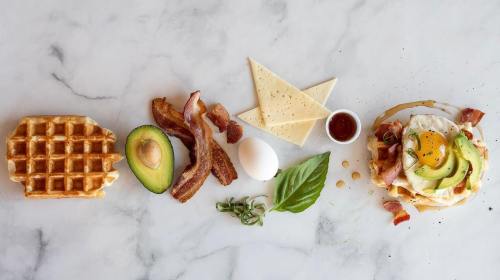 A spread of breakfast items: waffle, bacon, avocado, egg, cheese, and herbs, arranged on a marble surface.