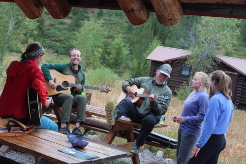 A group of people playing guitars and singing outdoors, surrounded by nature and rustic cabins.