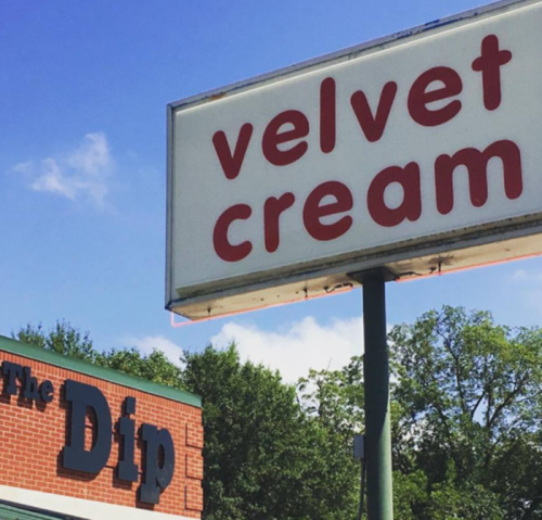Sign for "Velvet Cream" above a building labeled "The Dip," set against a blue sky and green trees.
