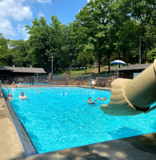 A sunny outdoor pool scene with people swimming and a water slide in the foreground, surrounded by trees.