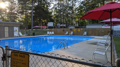 A fenced swimming pool with lounge chairs and umbrellas, surrounded by trees and a building in the background.