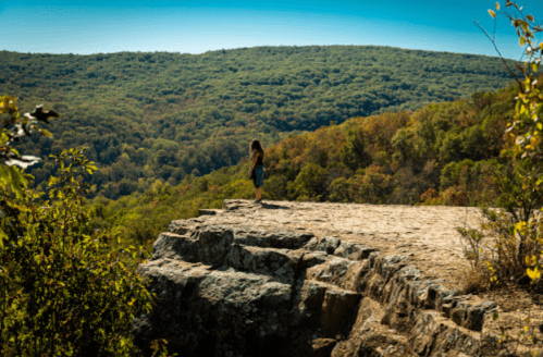 A person stands on a rocky ledge overlooking a lush, green valley under a clear blue sky.