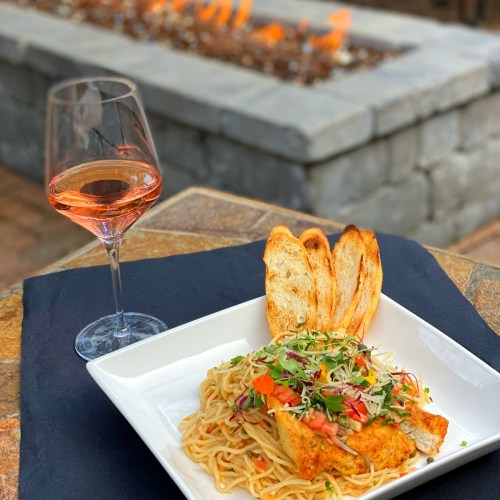 A plate of pasta topped with grilled chicken and vegetables, accompanied by toasted bread and a glass of rosé wine.