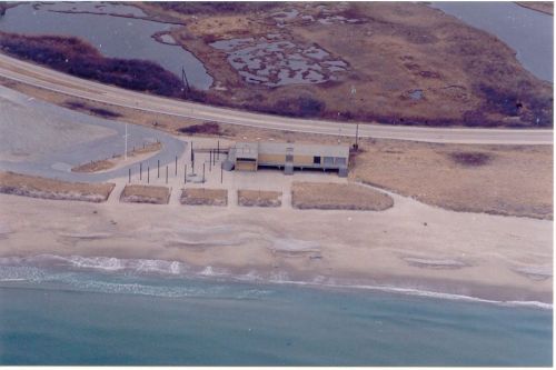Aerial view of a beach with a building, sandy shore, and a road nearby, surrounded by water and marshland.