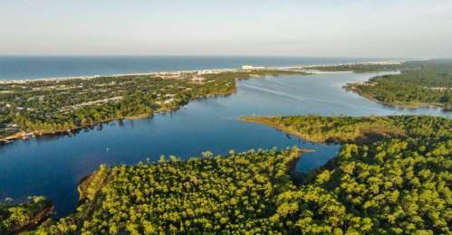 Aerial view of a lush green landscape with a river flowing into the ocean, surrounded by trees and coastal development.