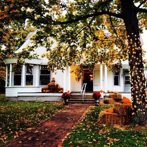 A charming white house decorated for fall, with pumpkins, hay bales, and lights on a tree amidst colorful autumn leaves.