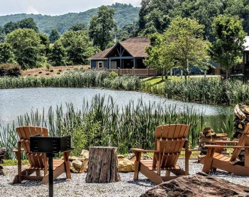 A serene lakeside scene with wooden chairs, a fire pit, and a house surrounded by greenery and mountains in the background.
