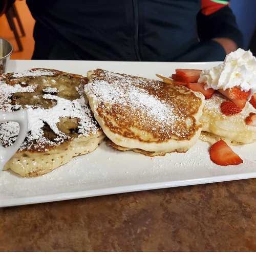 A plate of fluffy pancakes topped with powdered sugar, strawberries, and whipped cream, served with a small syrup cup.