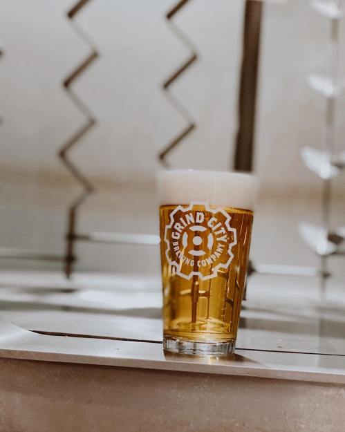 A glass of beer with a frothy head, featuring the Grind City Brewing Company logo, set against a modern background.