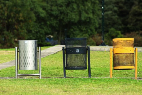 Three distinct trash bins in a grassy area, featuring silver, black, and yellow designs.