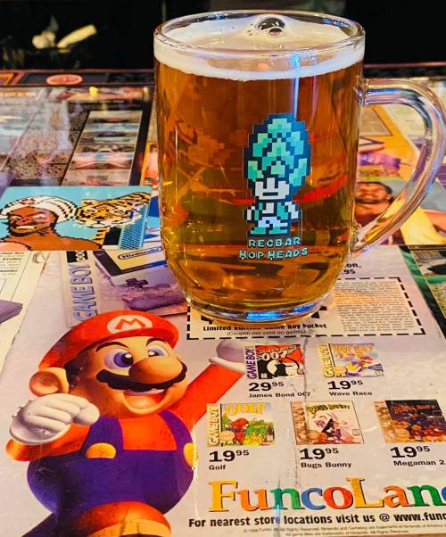 A glass of beer with a pixelated character design sits on a colorful arcade-themed table featuring Mario.