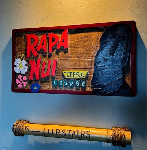 Sign for "Rapa Nui Tiki Lounge" featuring a stylized face and tropical flowers, with an arrow pointing "UPSTAIRS."