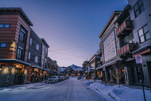 Snow-covered street lined with buildings, twinkling lights, and parked cars under a twilight sky.