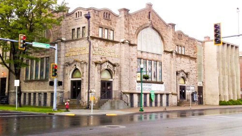 Historic stone building with large windows, featuring a sign for a Methodist church, set on a rainy street corner.
