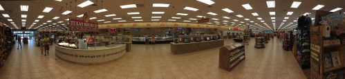 A spacious grocery store interior with aisles, displays, and a deli counter, featuring bright lighting and various products.