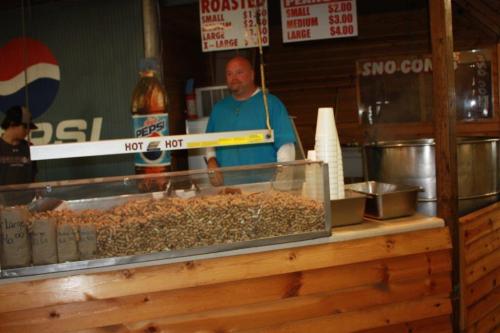 A man stands behind a counter filled with roasted peanuts, with Pepsi bottles and pricing signs visible in the background.