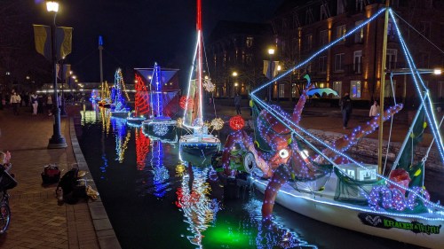 Colorful, illuminated boats decorated with lights reflect on a calm waterway at night, with people enjoying the scene.