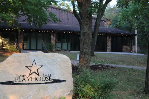 A stone sign for Timber Lake Playhouse in front of a building surrounded by trees and outdoor lighting.