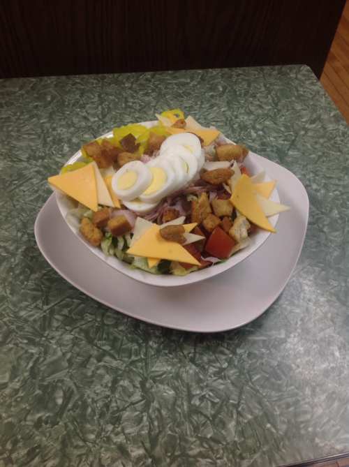 A colorful salad in a white bowl, topped with sliced eggs, cheese, croutons, and various vegetables on a green table.