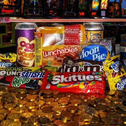 A colorful assortment of snacks including Lunchables, Skittles, and Moon Pie, set against a backdrop of arcade machines.