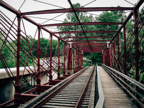 A red metal bridge with wooden planks and railway tracks, surrounded by green trees and a river.