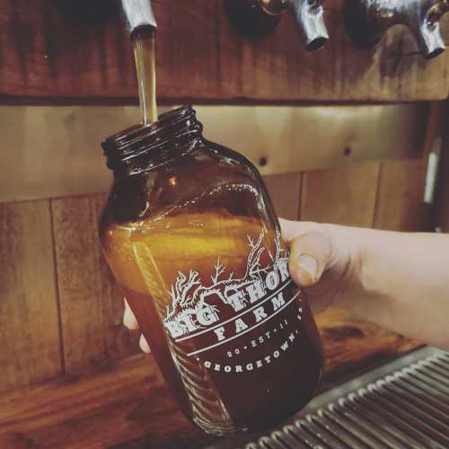 A hand pours a drink from a tap into a brown glass jar labeled "Big Thor Farm" against a wooden backdrop.
