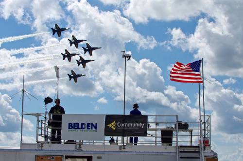 Aerial display of six jets flying in formation against a cloudy sky, with an American flag and spectators below.
