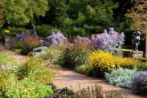 A vibrant garden path lined with colorful flowers and greenery, featuring statues among the lush landscape.