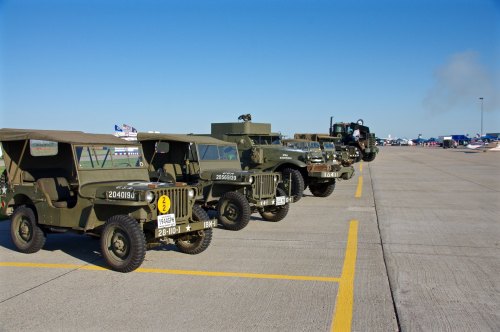 A row of vintage military vehicles parked on a tarmac under a clear blue sky.