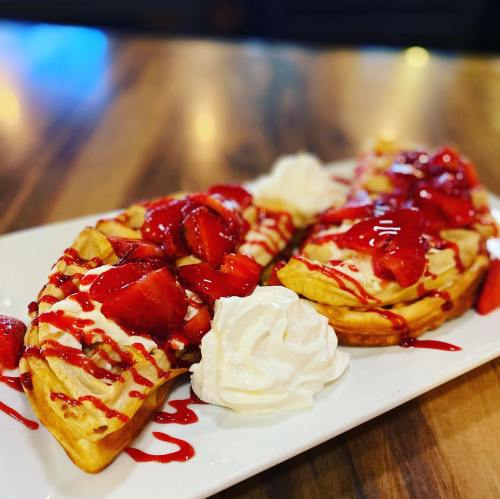 Two waffles topped with strawberries, whipped cream, and drizzled with strawberry sauce on a white plate.