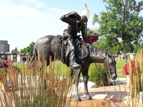 A bronze statue of a cowboy on horseback, tipping his hat, surrounded by greenery and flowers.